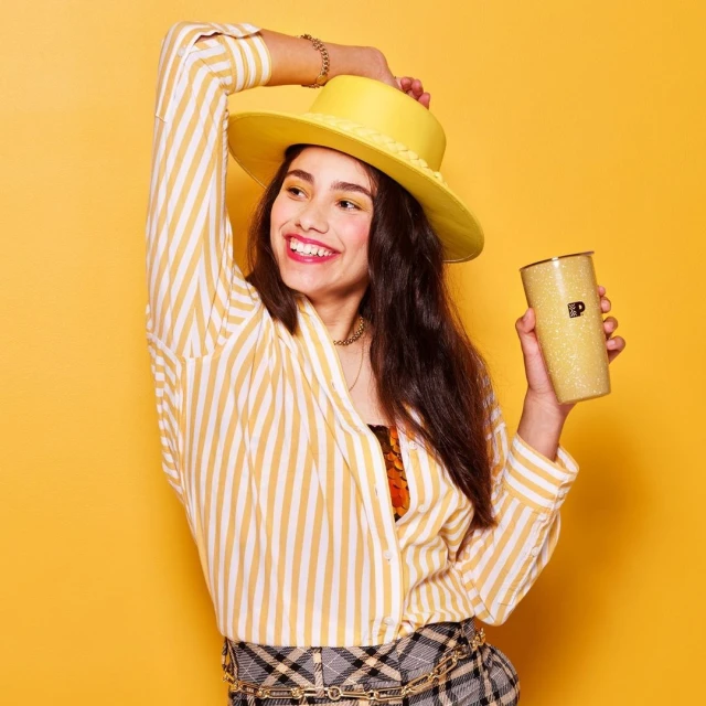 Young woman in striped shirt, hat, and plaid skirt, holding drink, smiles against a yellow backdrop in a vibrant, joyful studio shot.