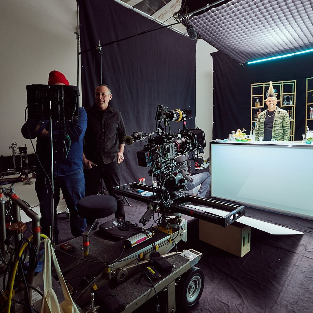 Behind-the-scenes shot of a film set with camera crew, lighting equipment, and a bartender preparing drinks in an indoor photo studio.