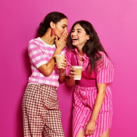 Two young women in pink outfits share a lighthearted moment with coffee cups against a bright pink backdrop, captured in a cheerful studio shot.