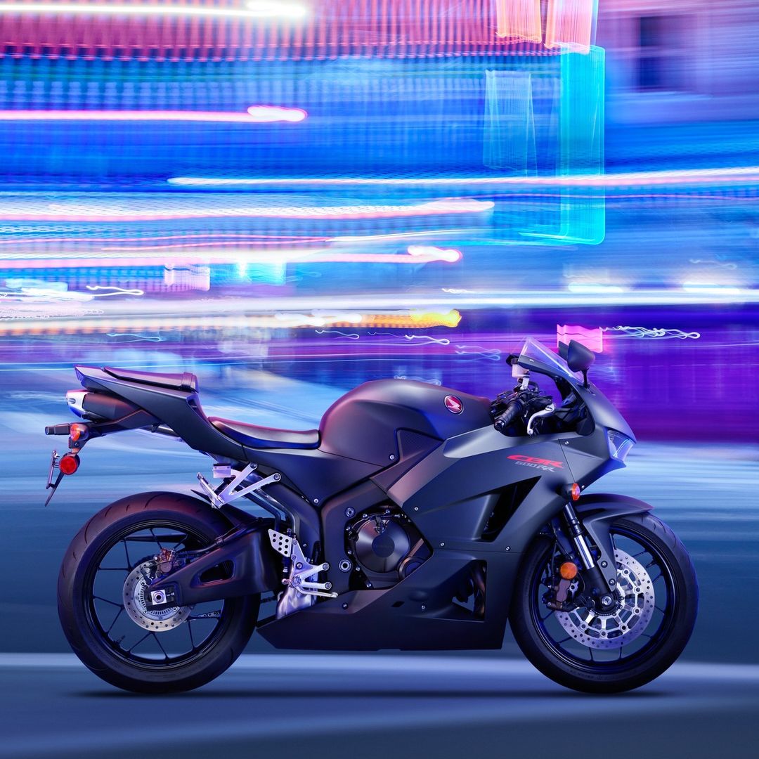 Black Honda CBR600RR motorcycle shown parked on a blue surface against an abstract background of neon blue, pink, and white light trails.