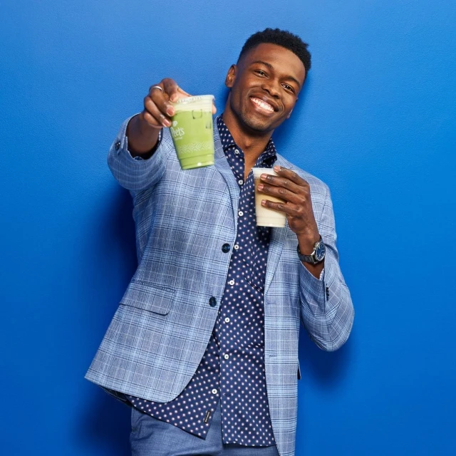 Smiling African-American man in a blazer holds up a green smoothie and a coffee cup in front of a bright blue background.