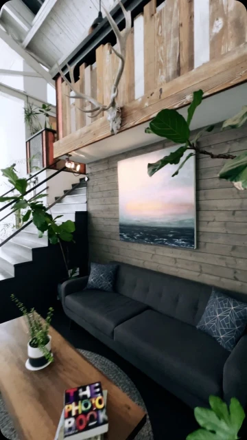 Living room interior featuring a grey sofa, coffee table with books, painting on a wood panel wall, and indoor plants.