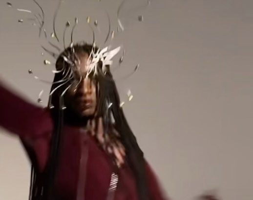 Portrait of a Black woman with an elaborate headpiece and a maroon-colored shirt against a grey background in a studio setting.