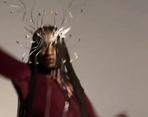 Portrait of a Black woman with an elaborate headpiece and a maroon-colored shirt against a grey background in a studio setting.