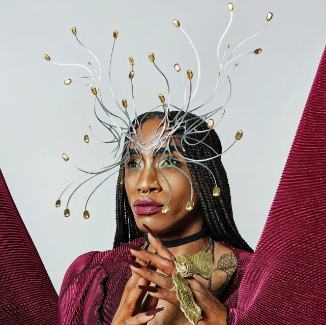Portrait of an androgynous person with unique metal headpiece and gold jewelry posing in front of a light gray background with red fabric.