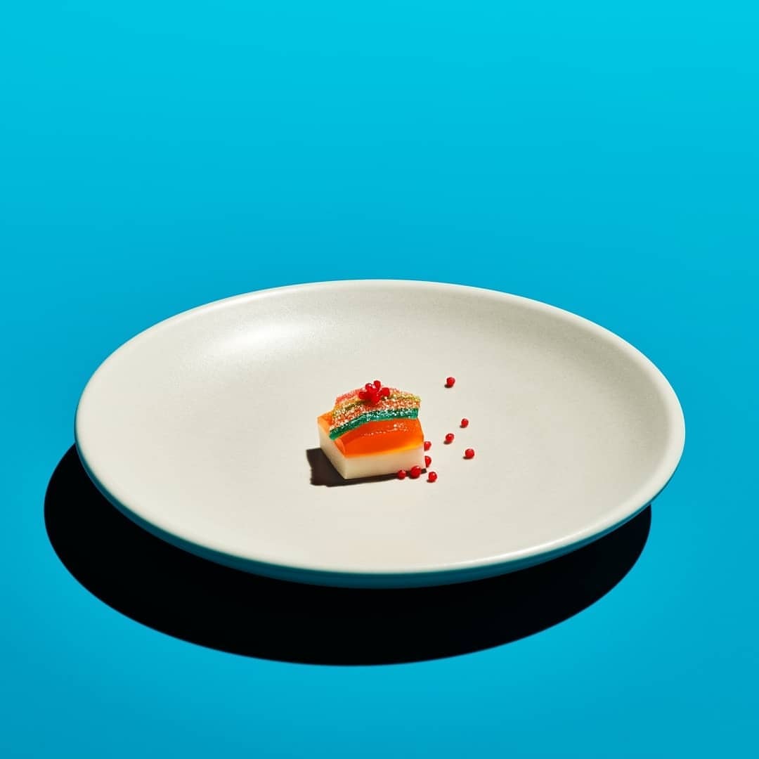 A piece of layered candy on a white plate is centered against a bright turquoise background, casting a shadow.