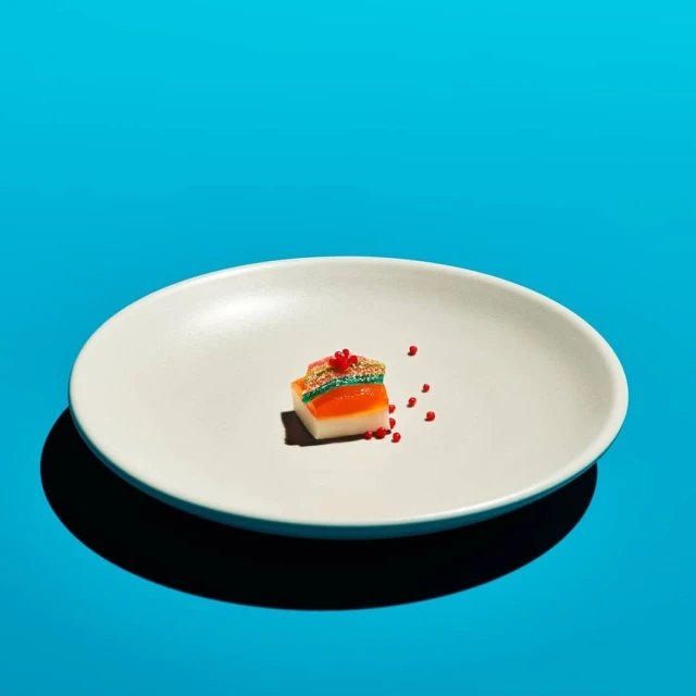 A piece of layered candy on a white plate is centered against a bright turquoise background, casting a shadow.