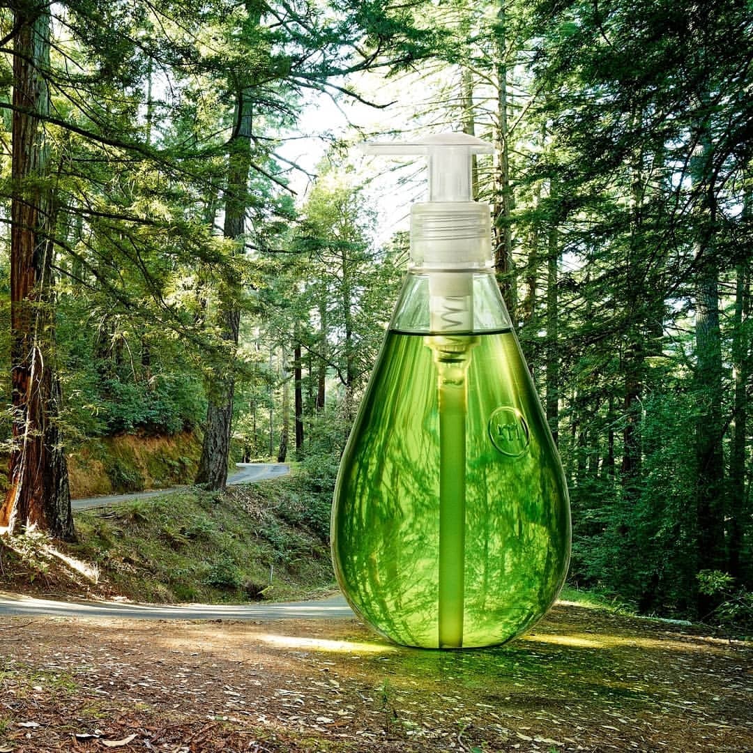 Green liquid soap in a clear dispenser stands in a forest. The product suggests cleanliness and harmony with nature.