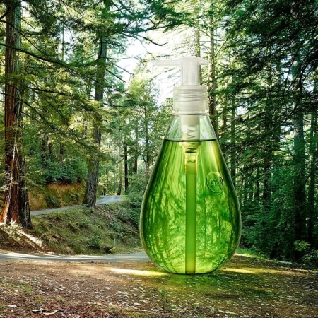Green liquid soap in a clear dispenser stands in a forest. The product suggests cleanliness and harmony with nature.