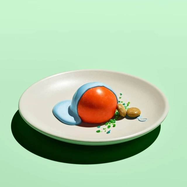Close-up of an orange sphere covered in blue liquid, presented on a white plate alongside peanuts and green sprinkles against mint backdrop.