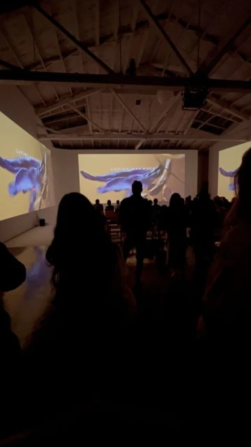 Indoor gathering of silhouetted people watching a projected video in a large room with bright overhead lighting and white walls.