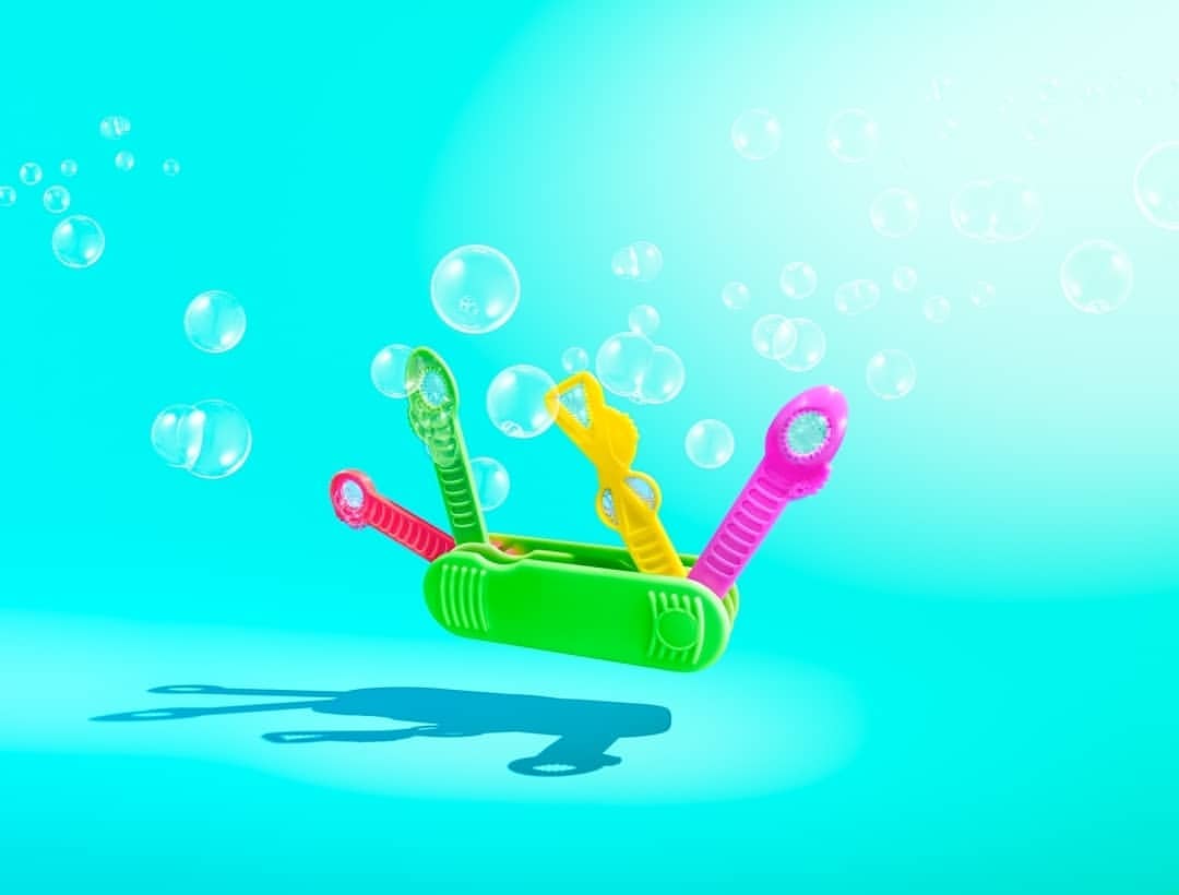 A green bubble wand with pink, yellow, and green attachments, sits against a turquoise backdrop surrounded by floating, transparent bubbles.