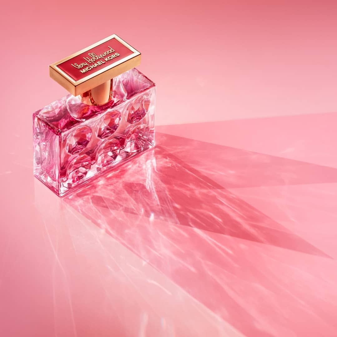 Rectangular perfume bottle with clear glass and a red label sits on a pink surface, casting a strong shadow in soft studio lighting.