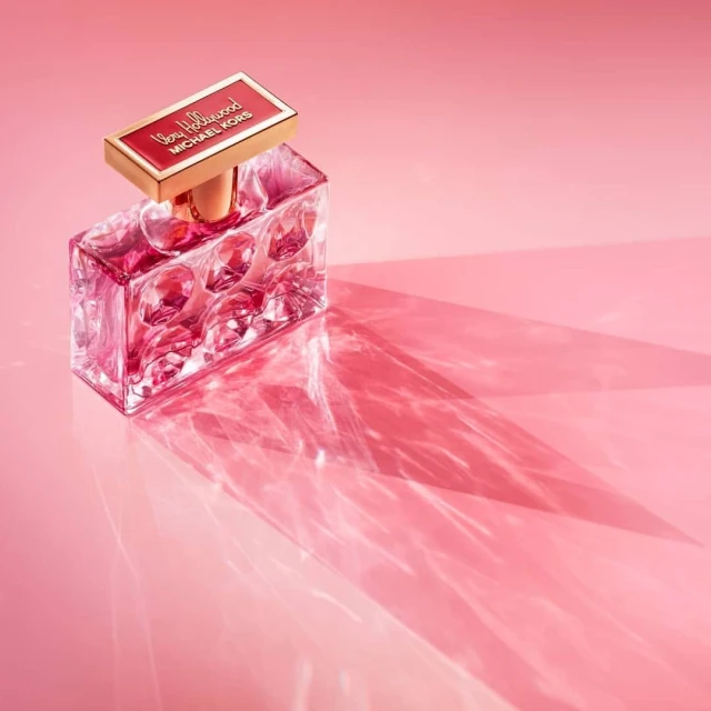 Rectangular perfume bottle with clear glass and a red label sits on a pink surface, casting a strong shadow in soft studio lighting.