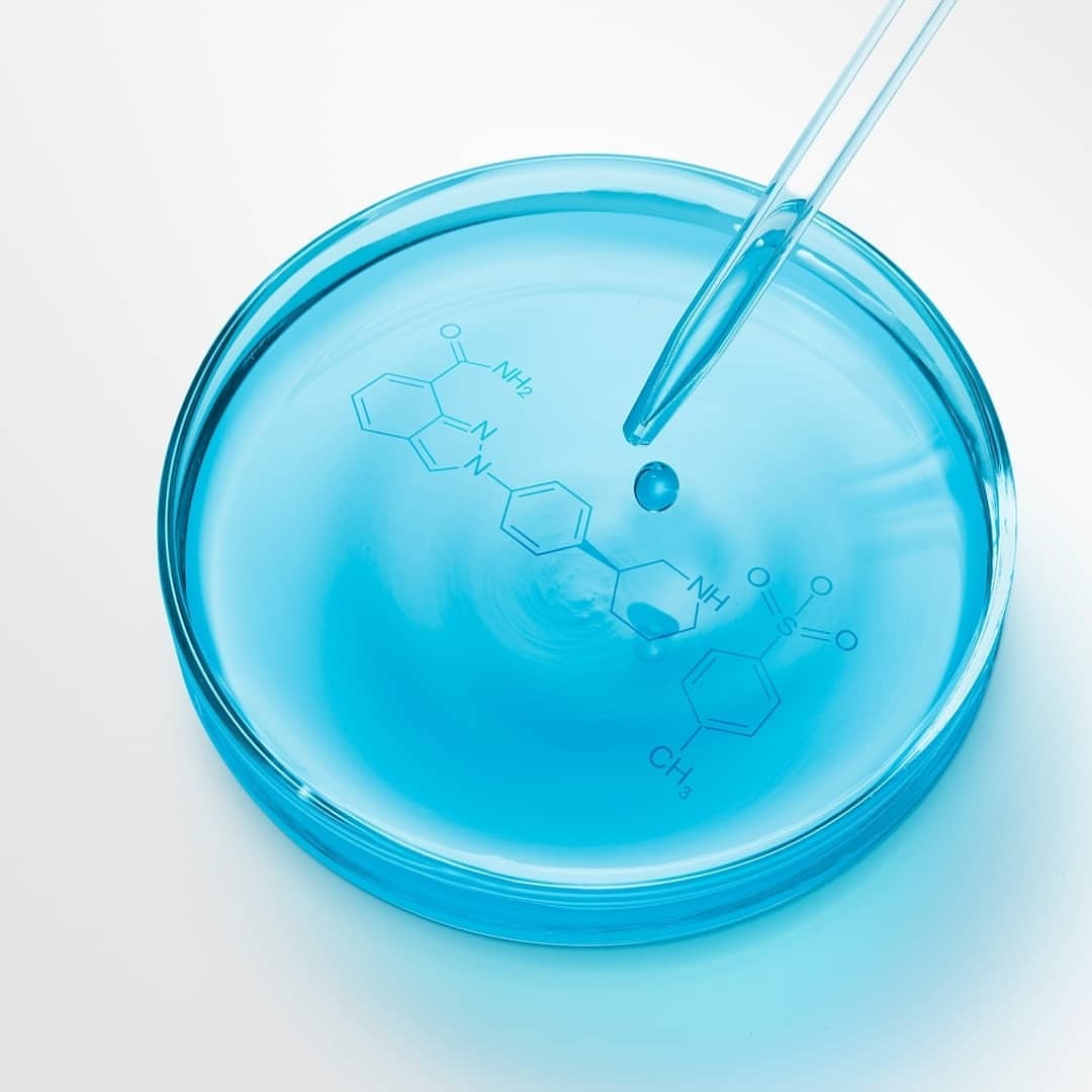 Clear pipette drips into a blue liquid-filled petri dish featuring a chemical structure diagram on a white surface in a studio setup.