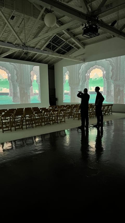 Two adult men stand in a large, brightly lit room with rows of wooden folding chairs facing projection screens displaying arched doorways.