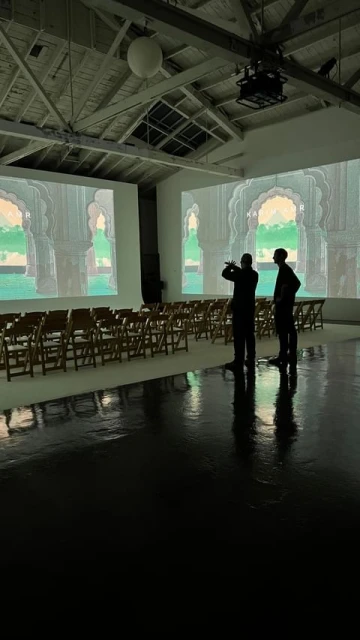 Two adult men stand in a large, brightly lit room with rows of wooden folding chairs facing projection screens displaying arched doorways.