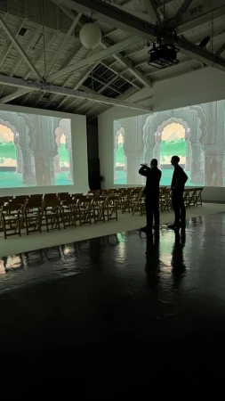 Two adult men stand in a large, brightly lit room with rows of wooden folding chairs facing projection screens displaying arched doorways.