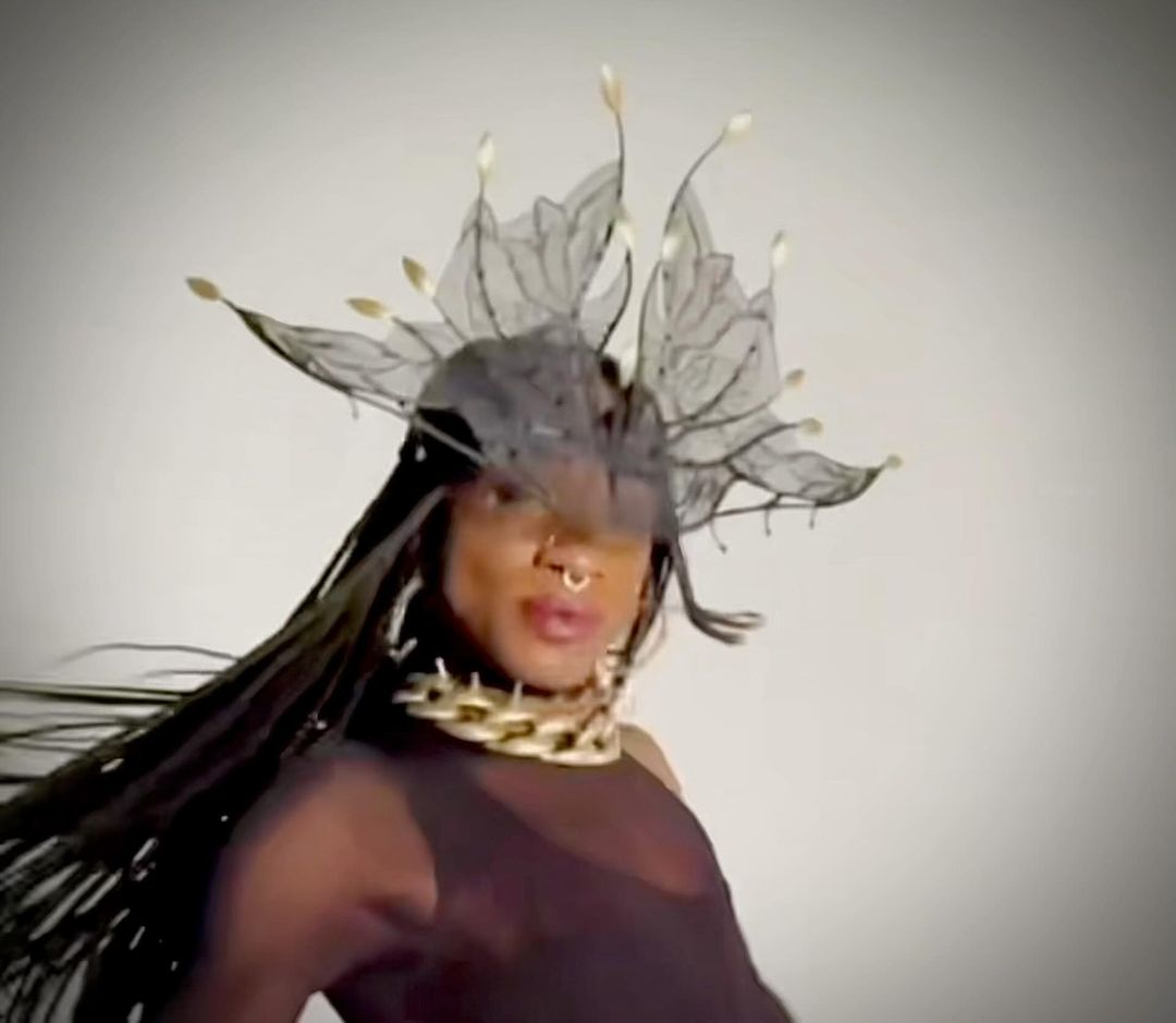 Stylish individual with dark hair and an elaborate headdress poses against a neutral background, wearing a gold chain and sheer top.