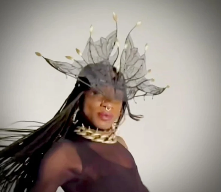 Stylish individual with dark hair and an elaborate headdress poses against a neutral background, wearing a gold chain and sheer top.