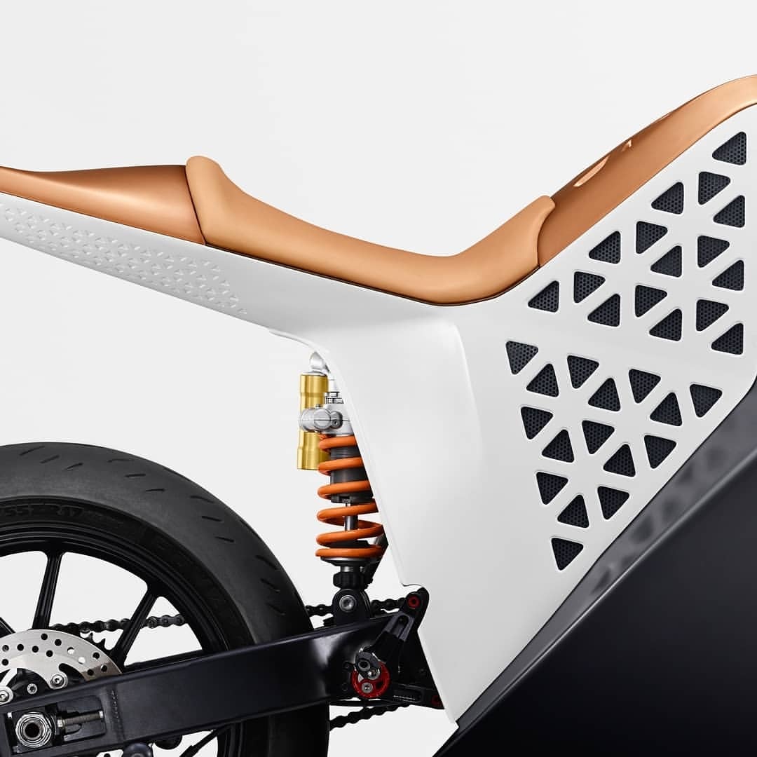 Close-up of a modern motorcycle featuring a white and tan frame, black tire, and orange shock absorber against a light background.