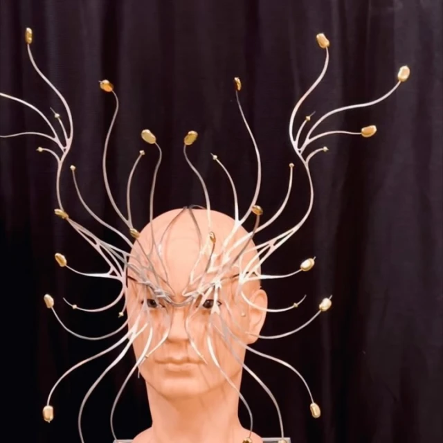 Headshot of a mannequin wearing an elaborate silver metal headpiece with gold accents against a dark background in a studio setting.