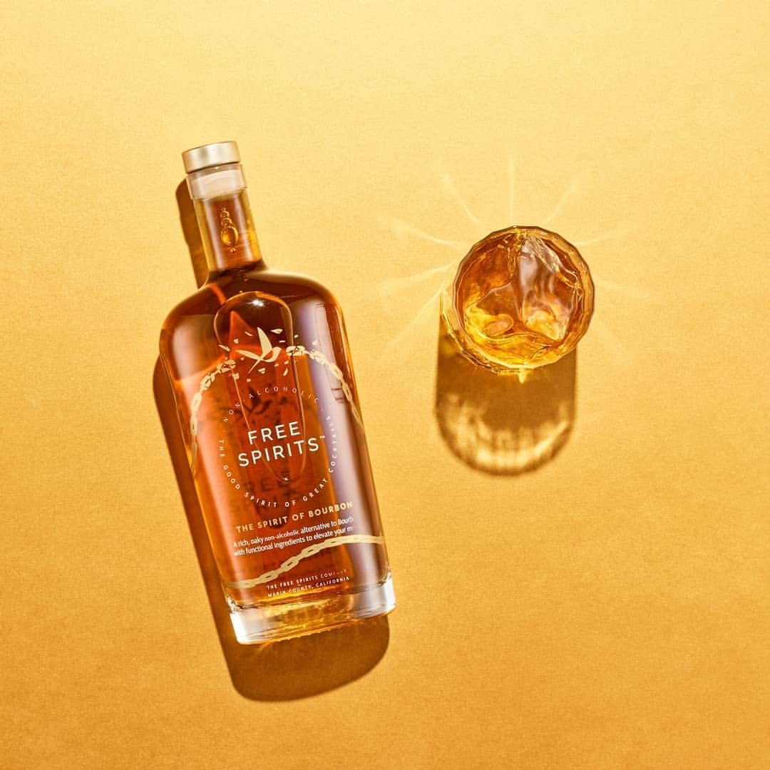 Bottle of Free Spirits bourbon-flavored non-alcoholic spirit with a glass of amber liquid on a yellow surface, studio lighting.
