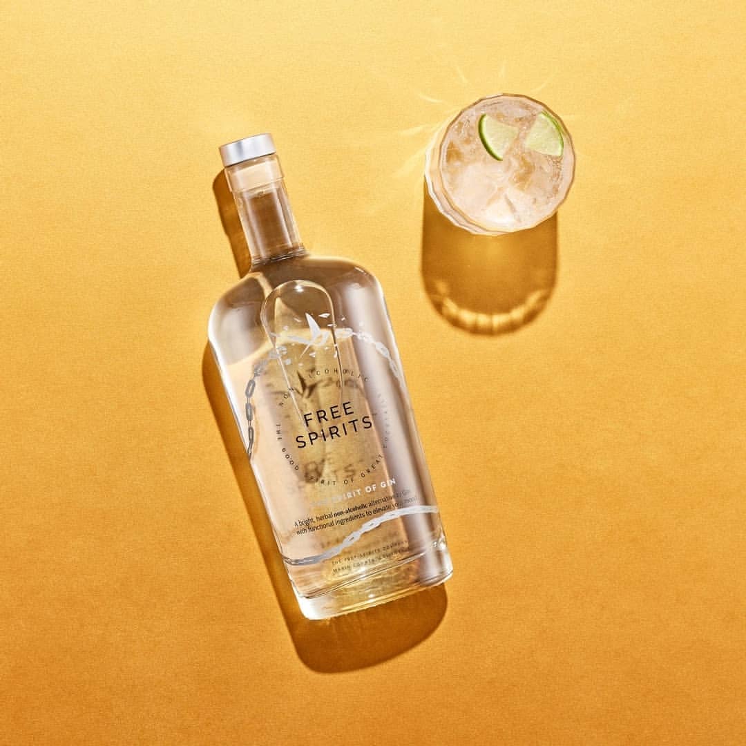 Studio shot of a clear glass bottle of gin and a glass with a gin cocktail and lime wedge, set against a warm golden background.