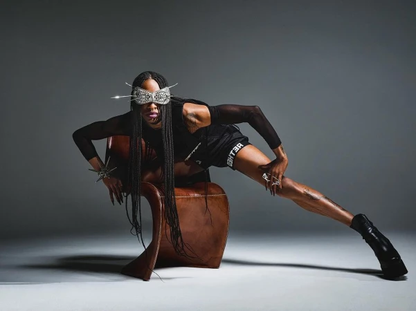 Model in black athletic shorts and long braids wearing an ornate bejeweled face mask, posing dramatically over a leather chair