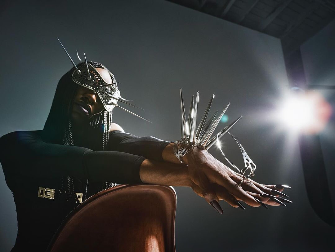 Model wearing avant-garde metallic spiky face mask and wrist armor resting arms on chair with bright lens flare in background.