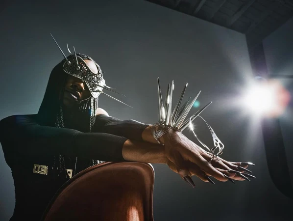 Model wearing avant-garde metallic spiky face mask and wrist armor resting arms on chair with bright lens flare in background.