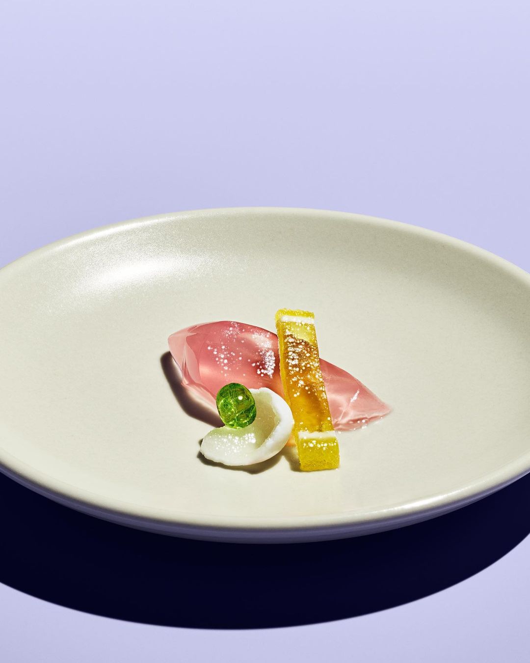 Close-up of food art on a white plate, featuring sorbet, fruit gel, and citrus candy, set against a light purple background.