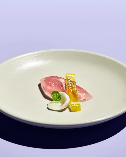 Close-up of food art on a white plate, featuring sorbet, fruit gel, and citrus candy, set against a light purple background.