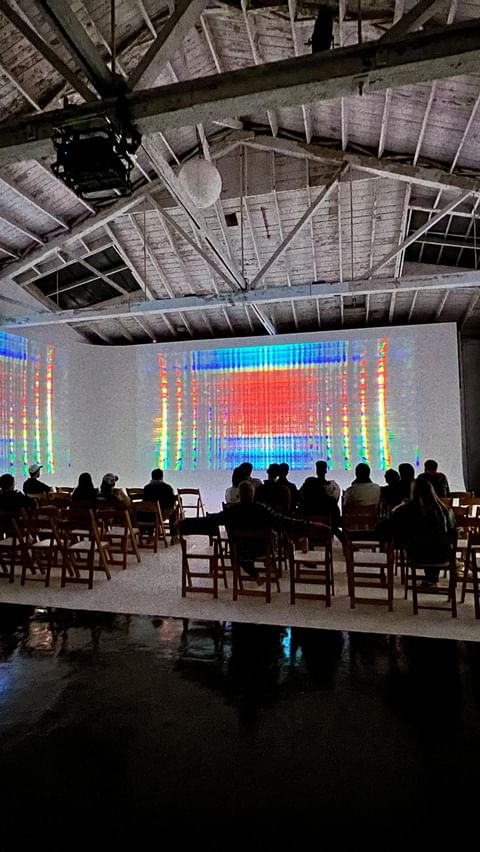 Audience sits in a warehouse facing a large screen displaying an abstract digital art piece with red, blue, and yellow colors.