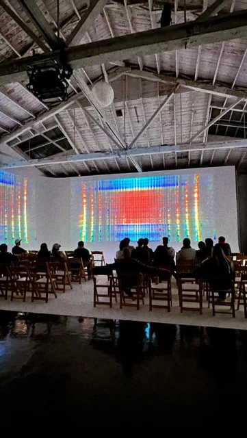 Audience sits in a warehouse facing a large screen displaying an abstract digital art piece with red, blue, and yellow colors.