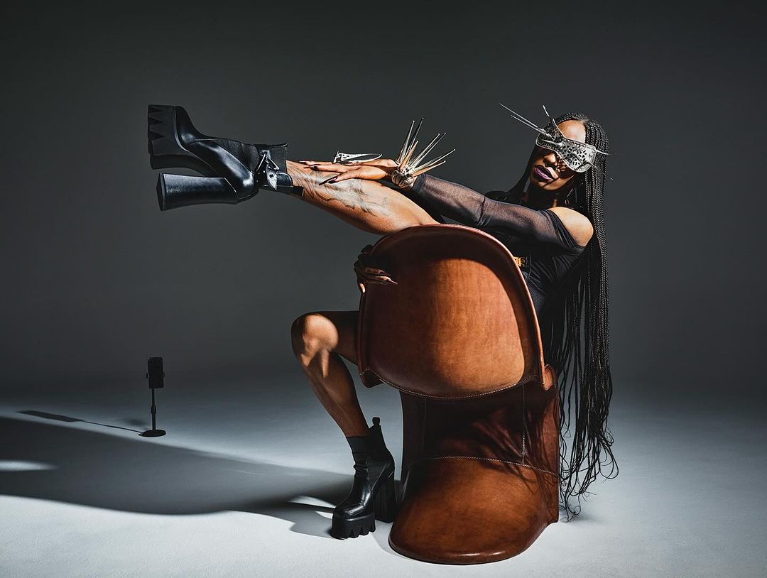 Person in avant-garde fashion sitting in a brown chair in a studio, wearing a silver mask, black top, and black platform boots.