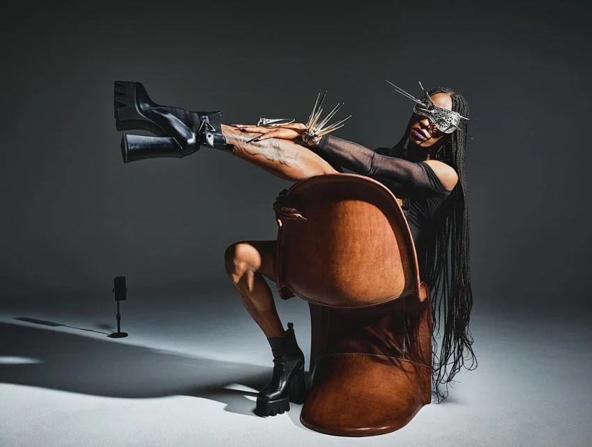 Person in avant-garde fashion sitting in a brown chair in a studio, wearing a silver mask, black top, and black platform boots.