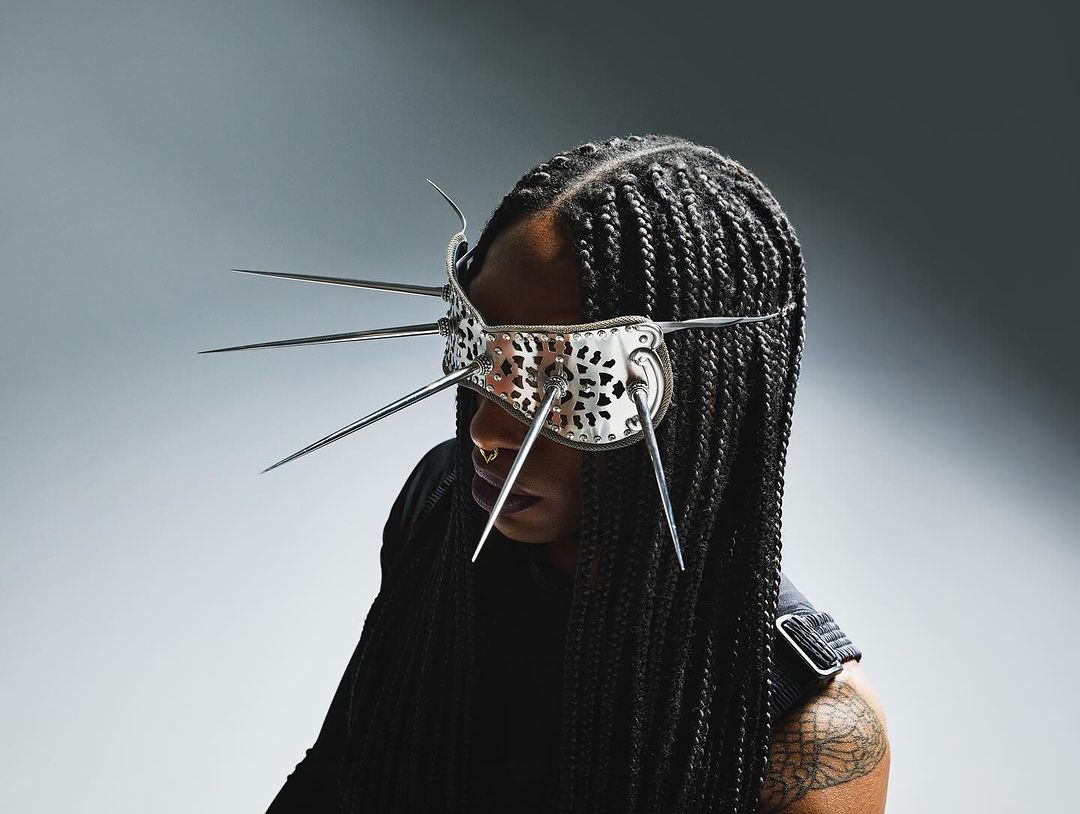 Woman with cornrow braids wearing a mirrored eye mask with spike details in a studio with even lighting against a gradient background.