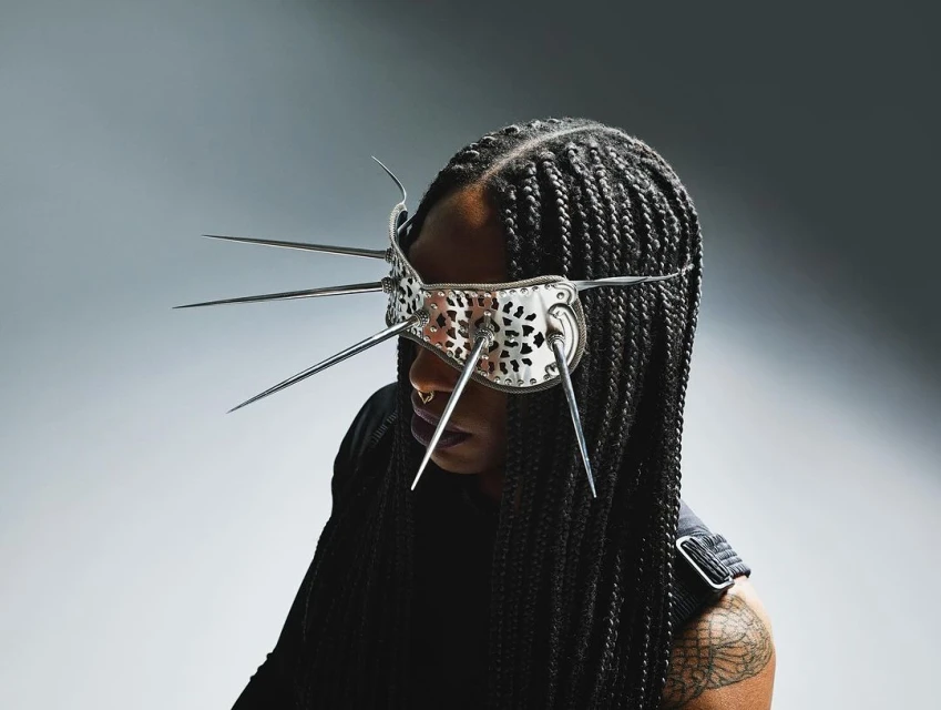 Woman with cornrow braids wearing a mirrored eye mask with spike details in a studio with even lighting against a gradient background.