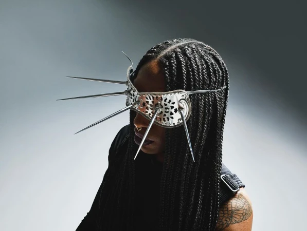 Woman with cornrow braids wearing a mirrored eye mask with spike details in a studio with even lighting against a gradient background.