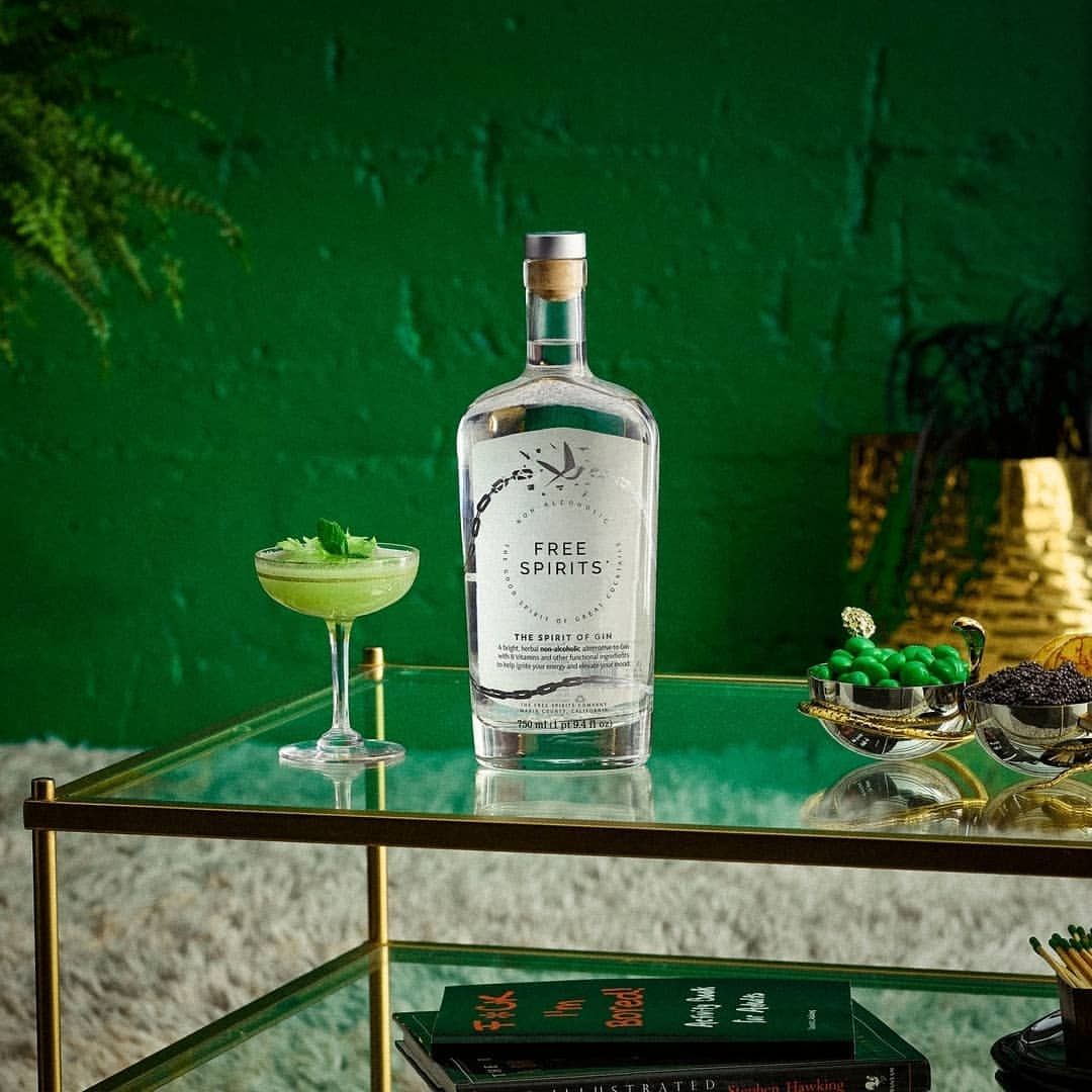 A clear gin bottle and a stemmed glass filled with a green drink sit on a glass coffee table with a green brick wall backdrop.