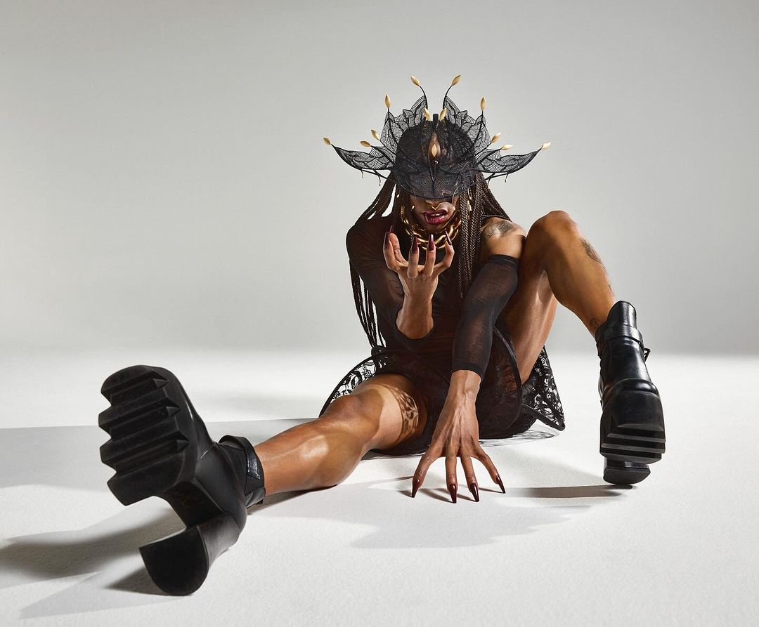 Person in elaborate lace mask and platform boots poses expressively in studio setting with neutral background. Conceptual fashion portrait.