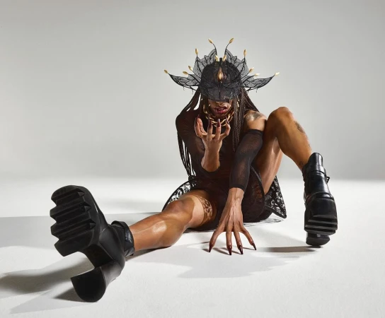 Person in elaborate lace mask and platform boots poses expressively in studio setting with neutral background. Conceptual fashion portrait.