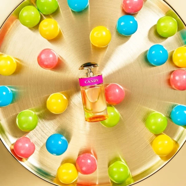 Perfume bottle on a gold plate surrounded by colorful gumballs, presented in a studio product shot with a bright and playful aesthetic.