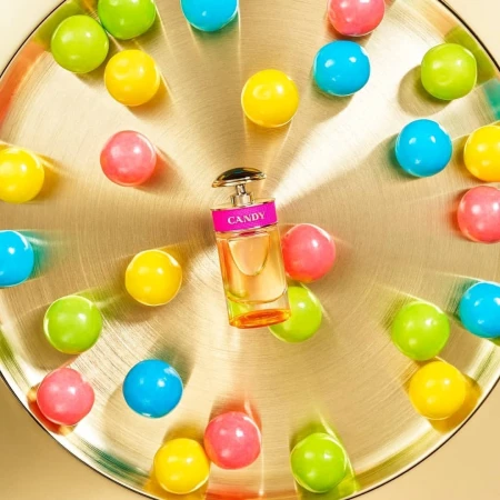 Perfume bottle on a gold plate surrounded by colorful gumballs, presented in a studio product shot with a bright and playful aesthetic.
