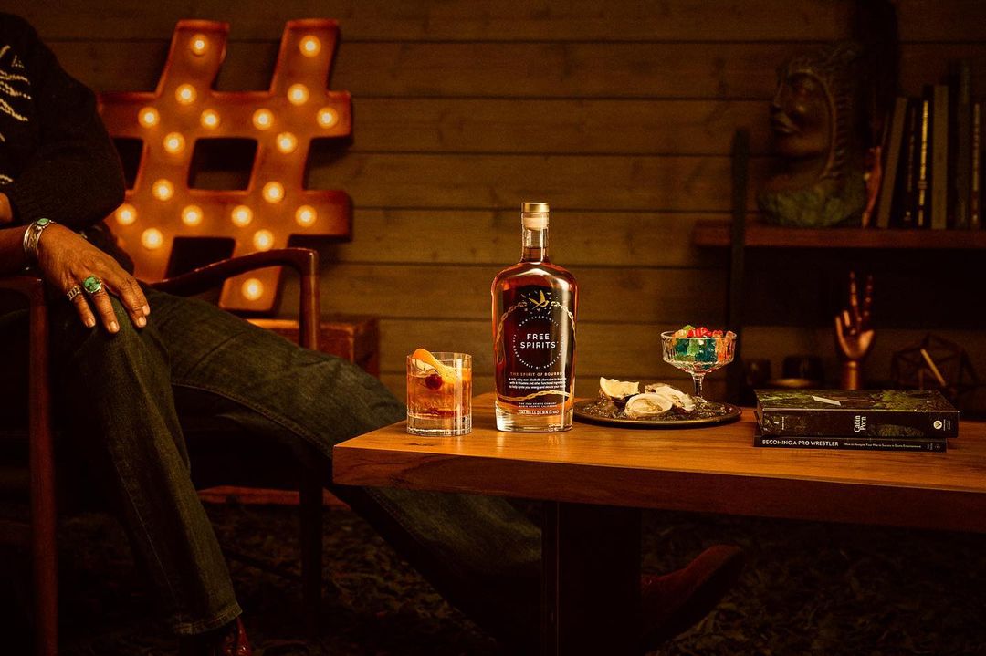 Bottle of Free Spirits stands on a wooden table with a cocktail, candies, and an adult wearing a bracelet and rings.