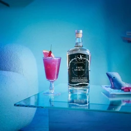 Glass bottle of liquor and a pink cocktail garnished with grapefruit and chili on a glass table in blue light