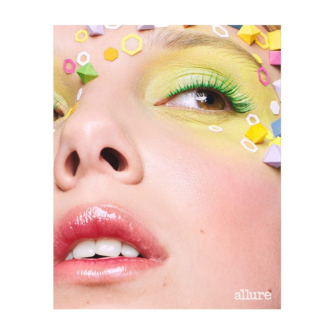 Close-up beauty shot of a young adult woman with bright yellow and green eyeshadow, long green eyelashes, and geometric paper art.