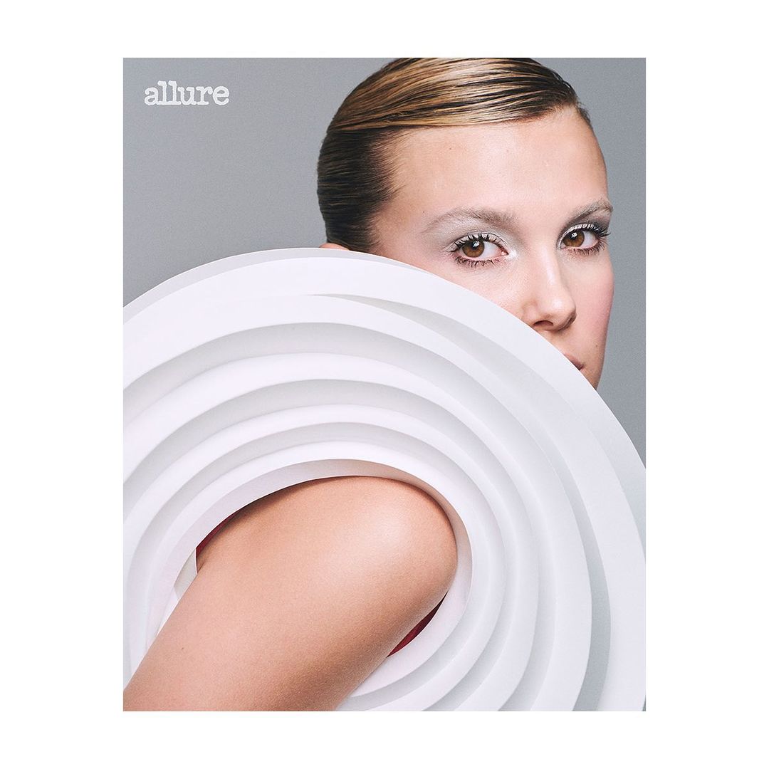 Close-up beauty shot of an adult female with slicked-back hair, pale skin, and light eyes, partially obscured by a white, tiered circular object.