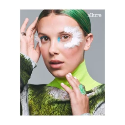 Close-up portrait of Millie Bobby Brown with green hair, white feathered makeup accents, and a bright neon green turtleneck.